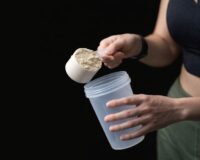 En person häller en skopa proteinpulver i en shakerflaska för träning.